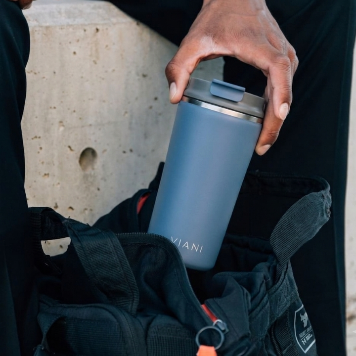 Vaso térmico MugsCafe VIANI azul sostenido en la mano junto a una mochila